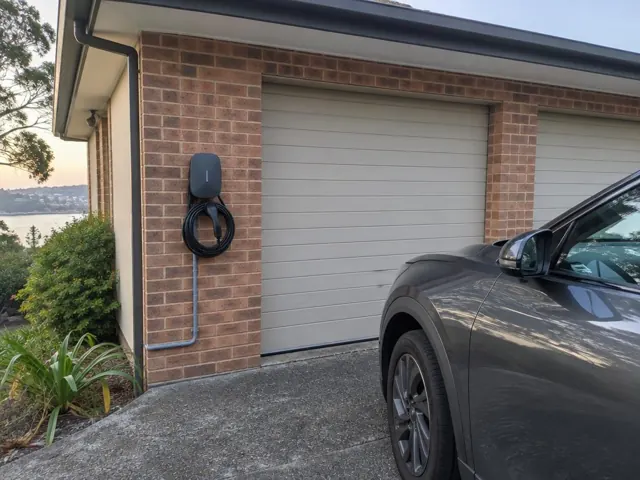 Wall-mounted EV charger and coiled cable on a brick garage wall beside a driveway in Chittaway Bay